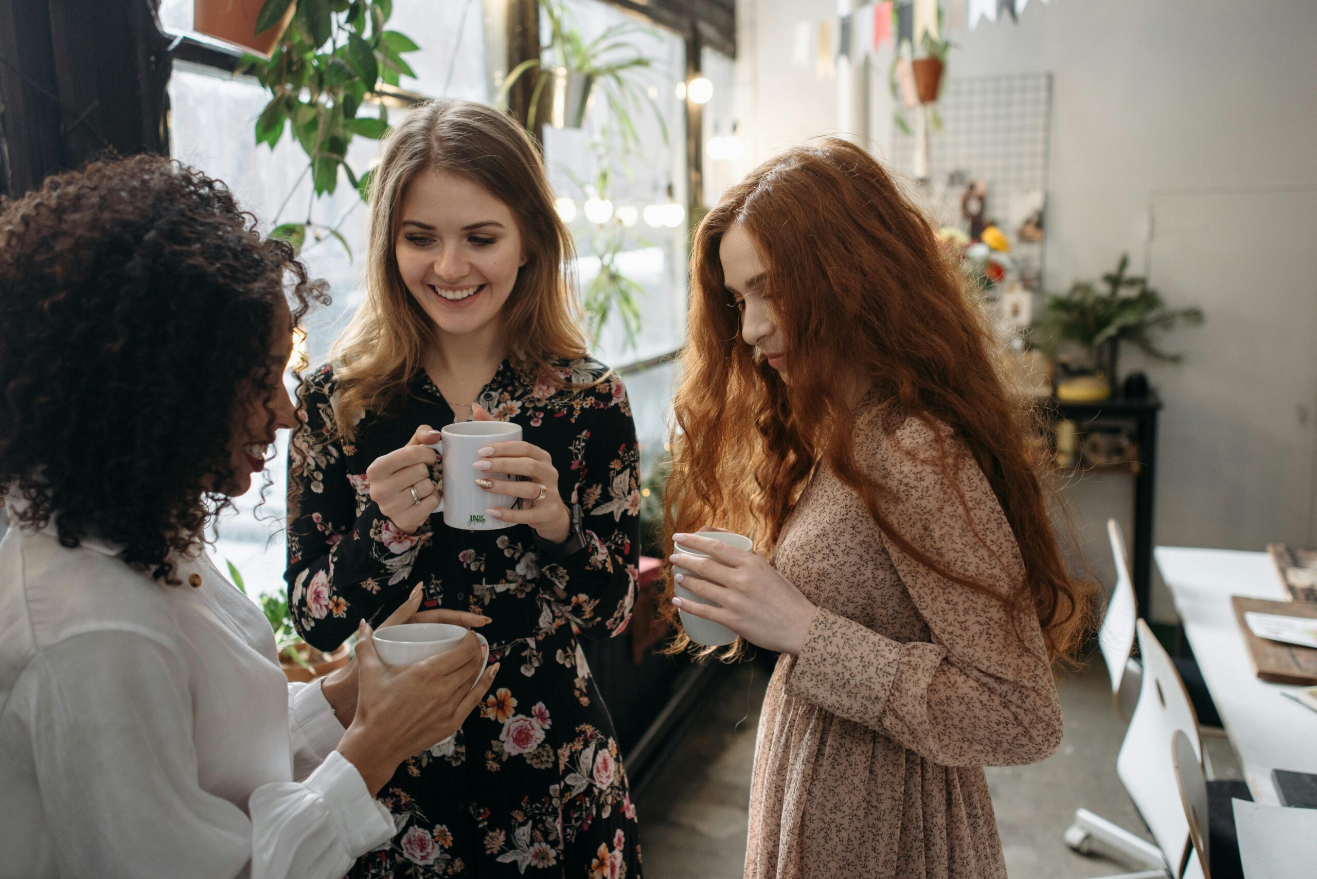 Three women standing together in a cozy space, smiling and holding coffee mugs, representing community and connection.